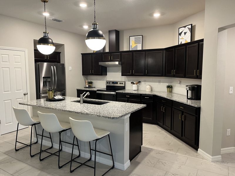 A modern kitchen with dark wood cabinetry, a sleek granite island, and stylish pendant lighting over white tile flooring.