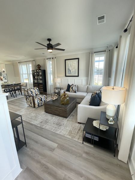 A bright living room with a neutral palette, featuring a plush sectional sofa, patterned accent chair, wooden coffee table, and soft grey curtains framing large windows.