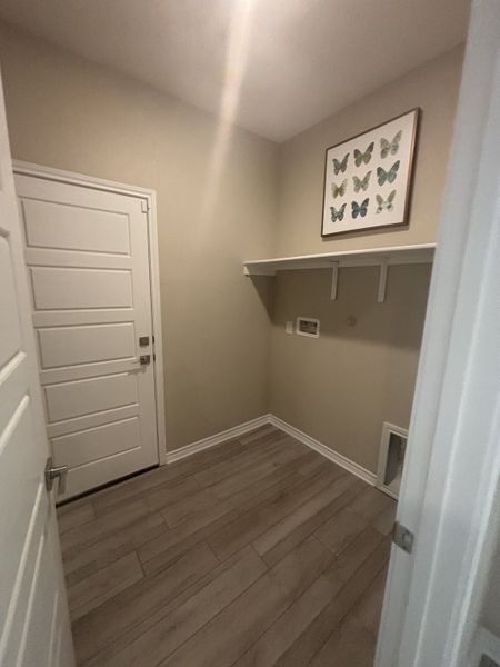 A cozy laundry room featuring wood-look tile flooring, creamy walls, a shelf, and butterfly artwork for a touch of charm. A cozy laundry room featuring wood-look tile flooring, creamy walls, a shelf, and butterfly artwork for a touch of charm.