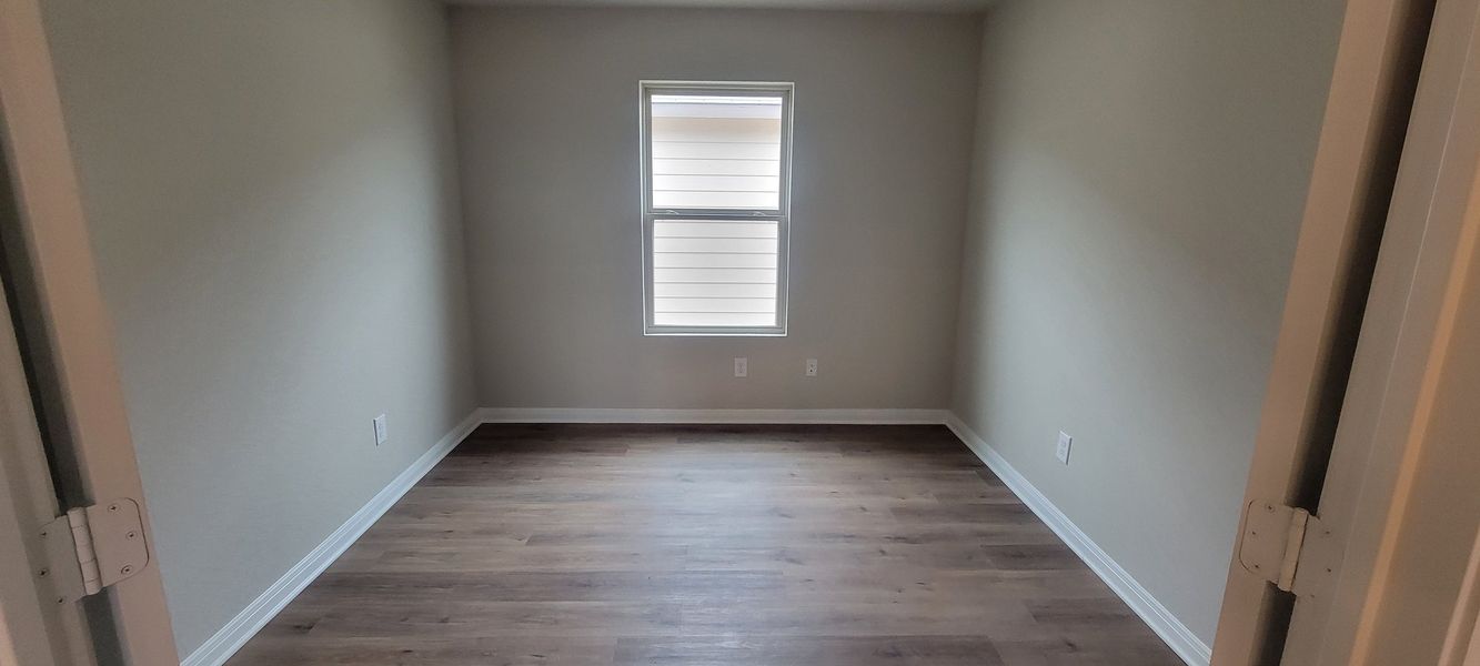 A bright room with soft gray walls, light wood flooring, and a single window offering natural light.