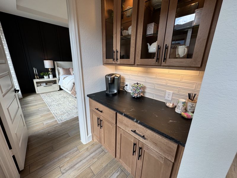 A cozy nook with wooden cabinets, black countertop, and coffee station leads to a chic bedroom with dark accent wall.
