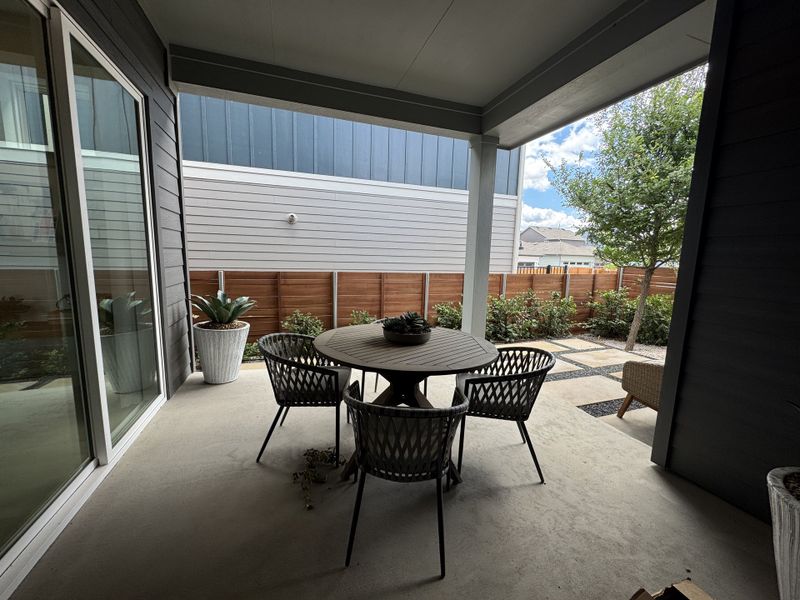 A cozy covered patio with a round table, modern chairs, potted plants, and a view of a landscaped yard.