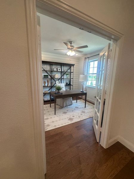 A cozy home office with a wooden desk, ceiling fan, and large window illuminating the space.