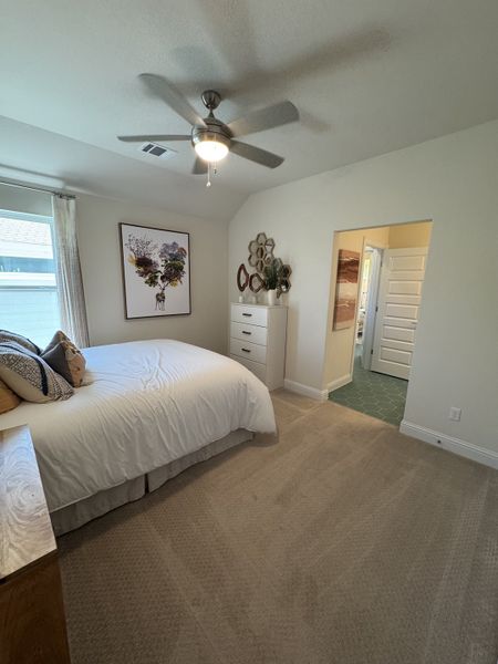 A cozy bedroom with soft carpeting, a ceiling fan, and tasteful decor, leading into a tiled bathroom.