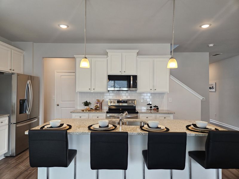 A modern kitchen with sleek black barstools, a granite island, pendant lighting, and stainless steel appliances.