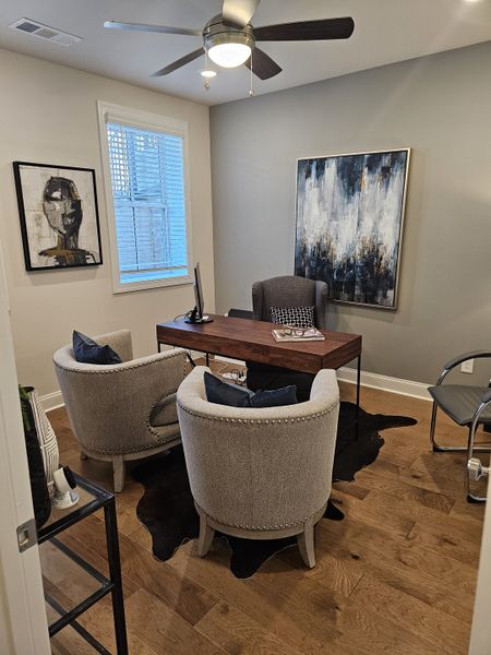 A cozy office with modern art, stylish seating, a wood desk, and contemporary ceiling fan, featuring elegant decor and hardwood floors.