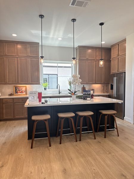 A modern kitchen with wooden cabinets, a sleek island, pendant lights, and bar stools on a warm wood floor.