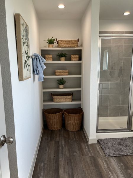 A cozy bathroom nook with woven baskets, plants, and wooden flooring, adjacent to a sleek glass shower.