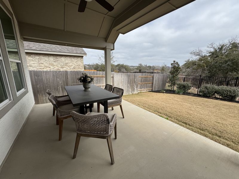 A cozy covered patio with a dining table and wicker chairs, overlooking a fenced backyard and landscaped garden.
