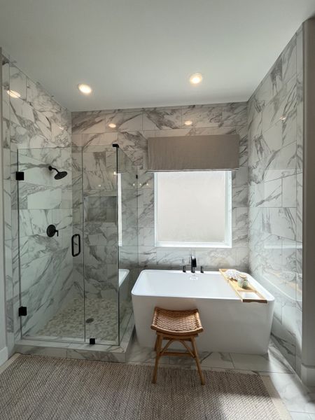 A luxurious bathroom with marble walls, a glass shower, elegant bathtub, and natural light streaming in.