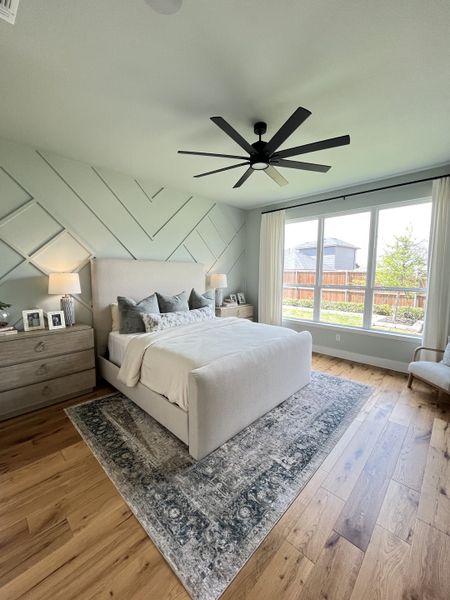 A serene bedroom with a plush bed, elegant paneling, large window, and stylish ceiling fan. Cozy and filled with natural light.