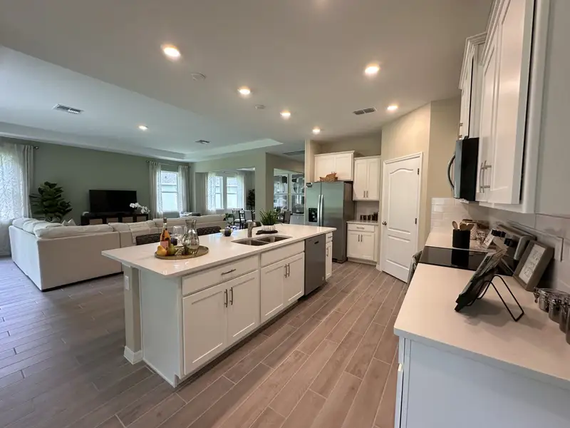 A spacious kitchen with white cabinetry, a central island, and an open layout leading to a cozy living area with large windows.