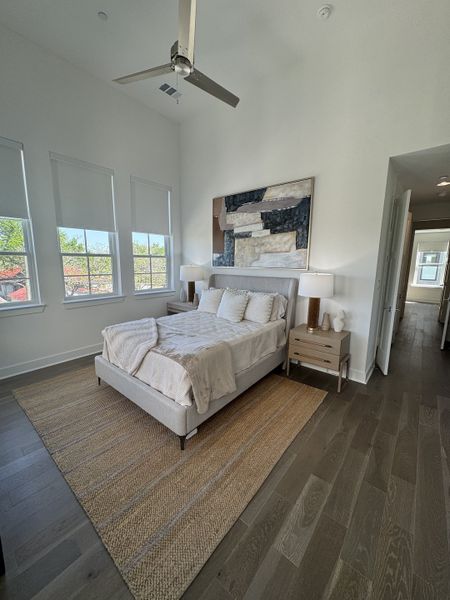 A cozy bedroom featuring a plush bed, neutral decor, artwork, and a stylish ceiling fan. Light streams in through large windows. A cozy bedroom featuring a plush bed, neutral decor, artwork, and a stylish ceiling fan. Light streams in through large windows.