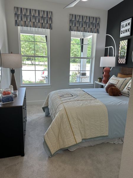A cozy bedroom featuring large windows, sporty decor, a soft carpet, and modern furnishings.