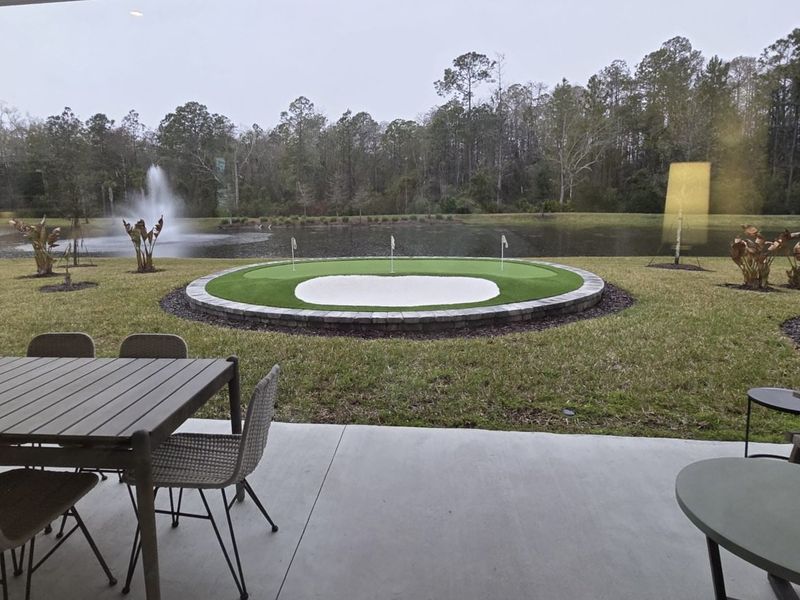 A serene patio view with a cozy seating area, putting green, and lake with a fountain in a tranquil setting. A serene patio view with a cozy seating area, putting green, and lake with a fountain in a tranquil setting.