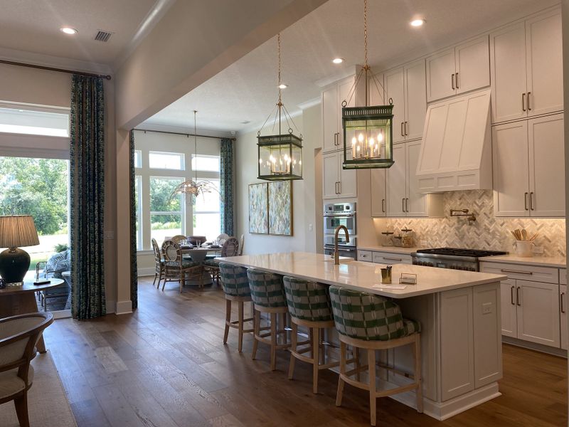 A spacious kitchen featuring a large island with elegant pendant lighting, white cabinets, and a dining area overlooking greenery.