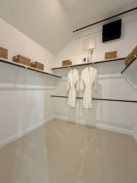 A spacious walk-in closet with dual hanging rails, beige carpet, and wicker storage baskets for organized living. A spacious walk-in closet with dual hanging rails, beige carpet, and wicker storage baskets for organized living.