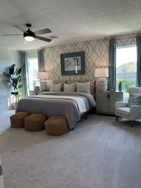 A cozy bedroom with patterned wallpaper, a large bed, wicker ottomans, and natural light from two windows.