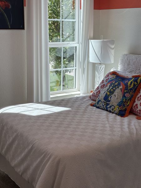 A cozy bedroom with a bright window, decorative pillows, and a soft white bedspread, creating a peaceful atmosphere.