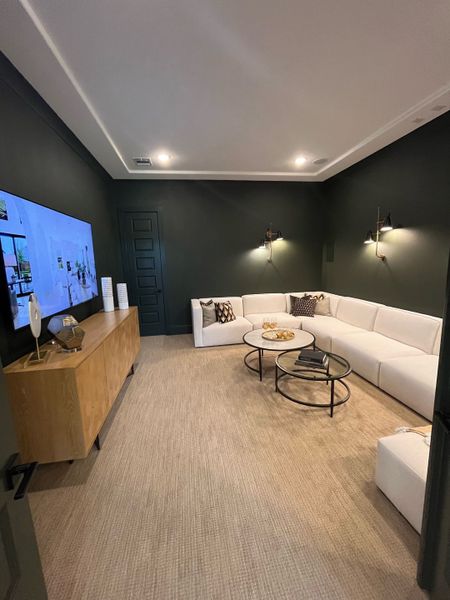 A cozy media room with dark walls, a large white sectional sofa, modern lighting, and a sleek wooden cabinet.