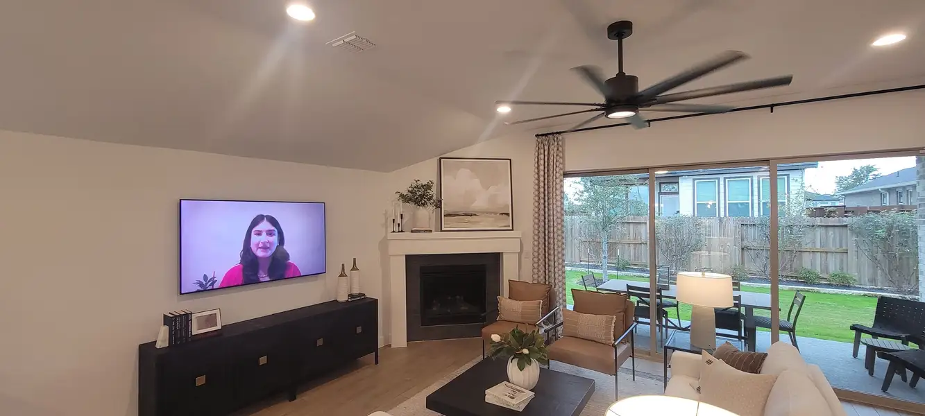 A cozy living room with a modern fireplace, large TV, and plush seating, overlooking a lush backyard through sliding glass doors.