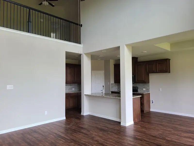 An open-concept kitchen with wooden cabinets, granite countertops, and dark wood flooring beneath a loft, creating airy elegance.