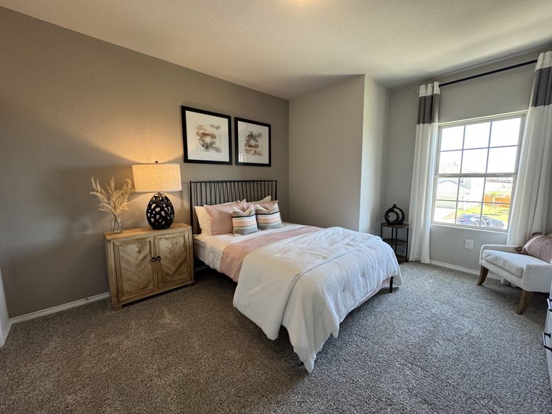 A cozy bedroom featuring a plush carpet, elegant wall art, natural wood nightstand, and a large window with stylish curtains.