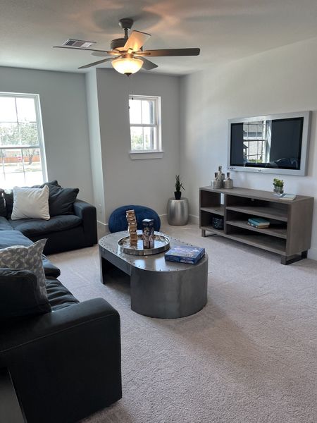A cozy living room featuring a sleek metallic coffee table, comfy seating, soft carpeting, and modern ceiling fan.