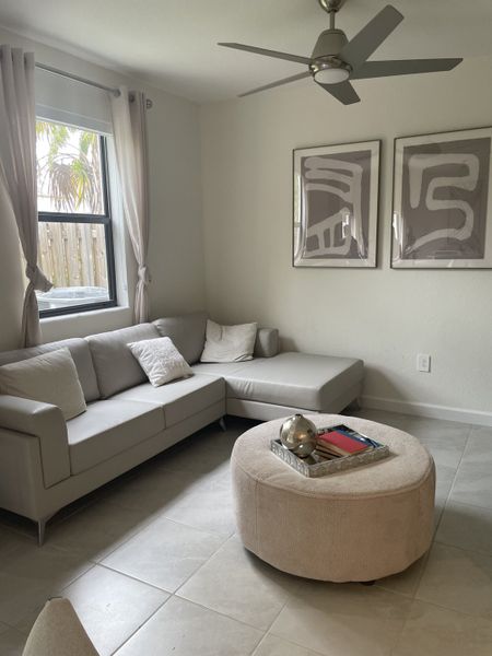 A cozy living room with a beige sectional sofa, modern art, tiled floors, and a ceiling fan, offering a serene ambiance.