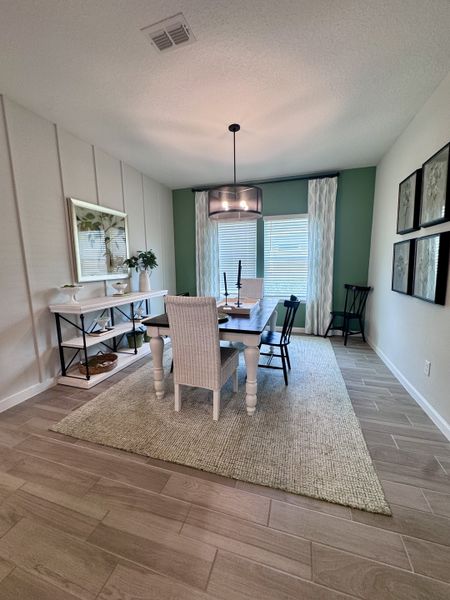 A cozy dining room with a rustic table, wicker chair, and modern lighting, accented by soft green walls and stylish decor.