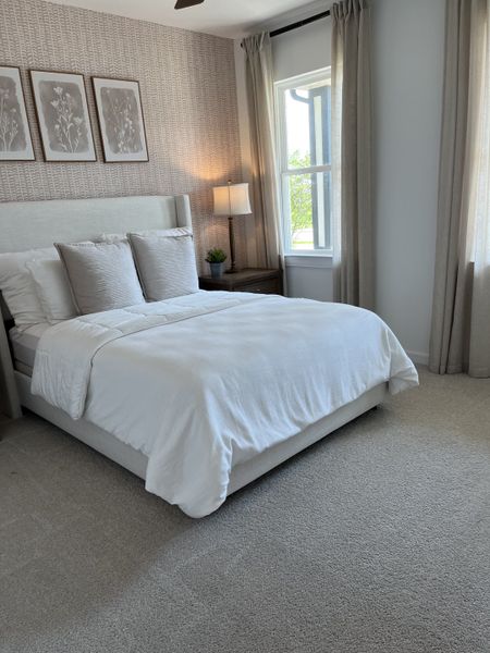 A cozy bedroom with soft carpeting, a plush bed, elegant wallpaper, and natural light filtering through large windows.