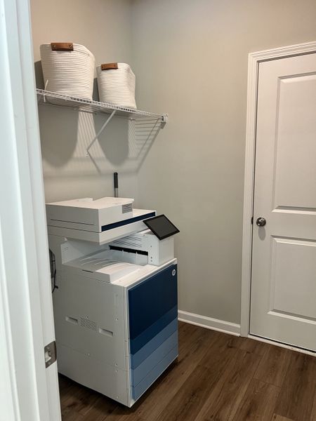 A functional utility room featuring a modern copier, wooden floors, and white storage baskets on a wire shelf.