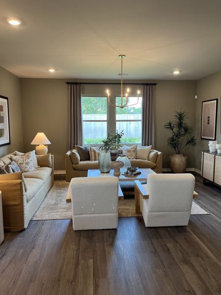 A cozy living room with plush seating, wood flooring, and a chic chandelier, bathed in natural light from large windows.
