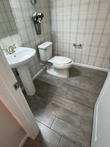 A chic bathroom featuring plaid wallpaper, a pedestal sink, and modern grey tiled flooring. A chic bathroom featuring plaid wallpaper, a pedestal sink, and modern grey tiled flooring.