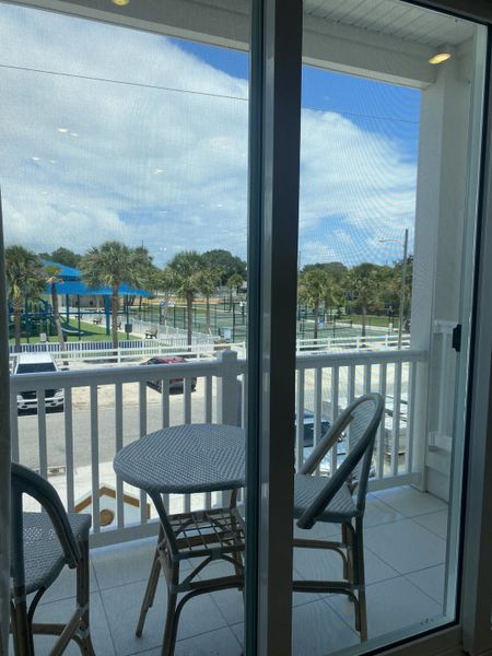 A cozy balcony with wicker chairs and a small table, offering a view of lush palm trees and a playground beyond. A cozy balcony with wicker chairs and a small table, offering a view of lush palm trees and a playground beyond.