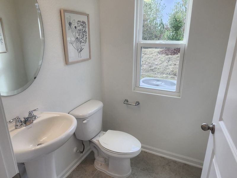 A bright bathroom with a pedestal sink, elegant mirror, floral art, and a window view.