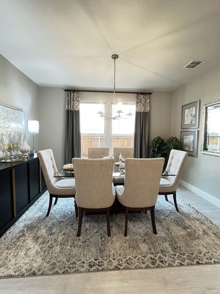 A cozy dining area with a glass table, beige upholstered chairs, elegant chandelier, and large windows with stylish curtains.