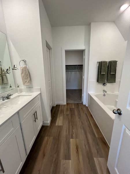 A modern bathroom with sleek wood flooring, dual sinks, and a spacious tub leading to a walk-in closet.