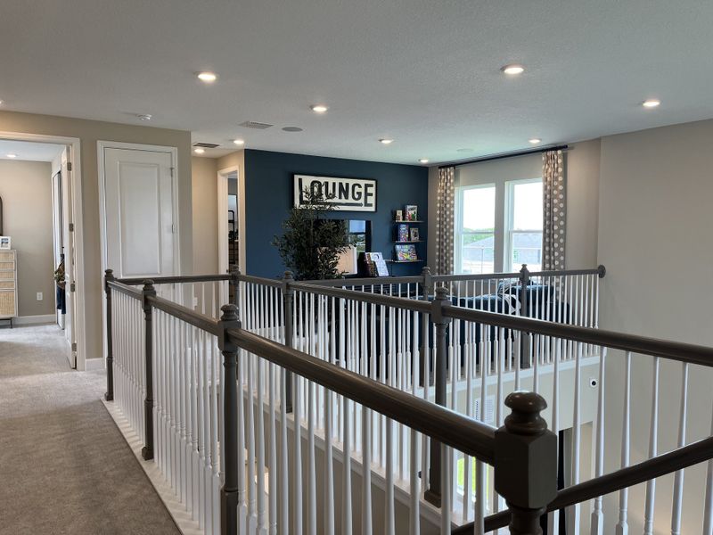 A cozy loft area with a plush carpet, blue accent wall, and lounge sign, overlooking a window-lit sitting space.