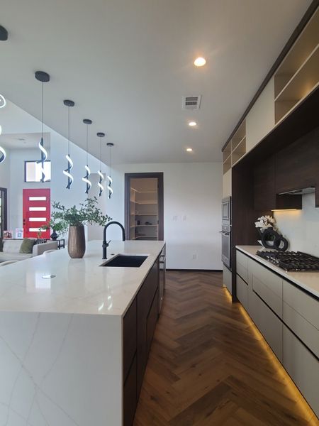 A modern kitchen with a sleek marble island, contemporary lighting, dark cabinets, and chevron wood flooring.