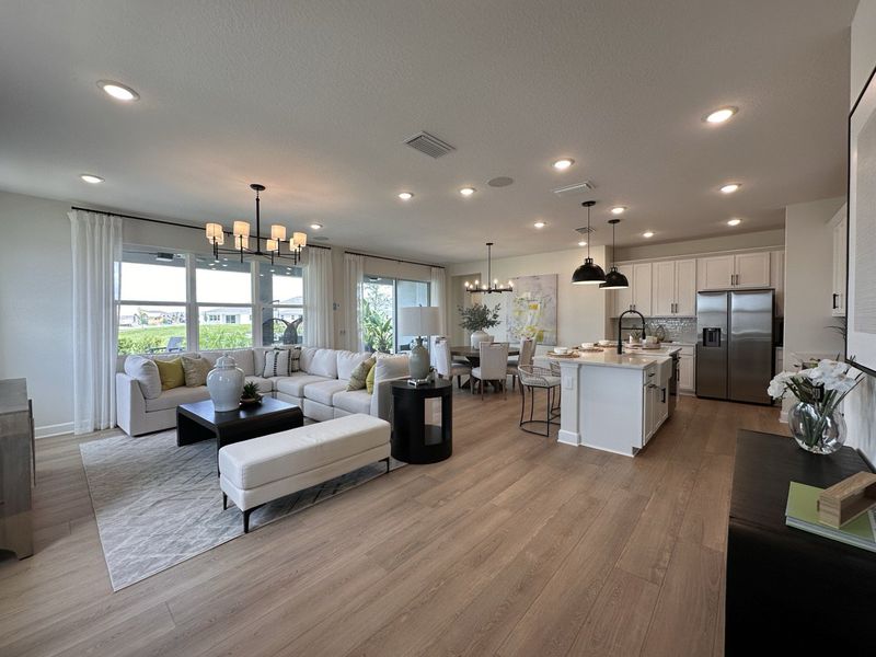 An open-concept living area featuring light wood floors, a modern kitchen, and elegant lighting with large windows.