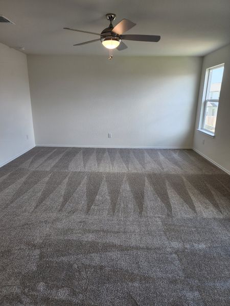 A spacious room with plush carpeting and a modern ceiling fan, bathed in natural light from a large window.