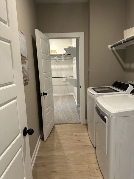 A functional laundry room with modern washer and dryer, light wood floors, and access to a walk-in closet.