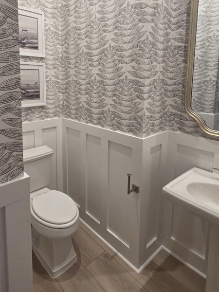 A charming bathroom with leaf-patterned wallpaper, wainscoting, and a sleek pedestal sink. A charming bathroom with leaf-patterned wallpaper, wainscoting, and a sleek pedestal sink.