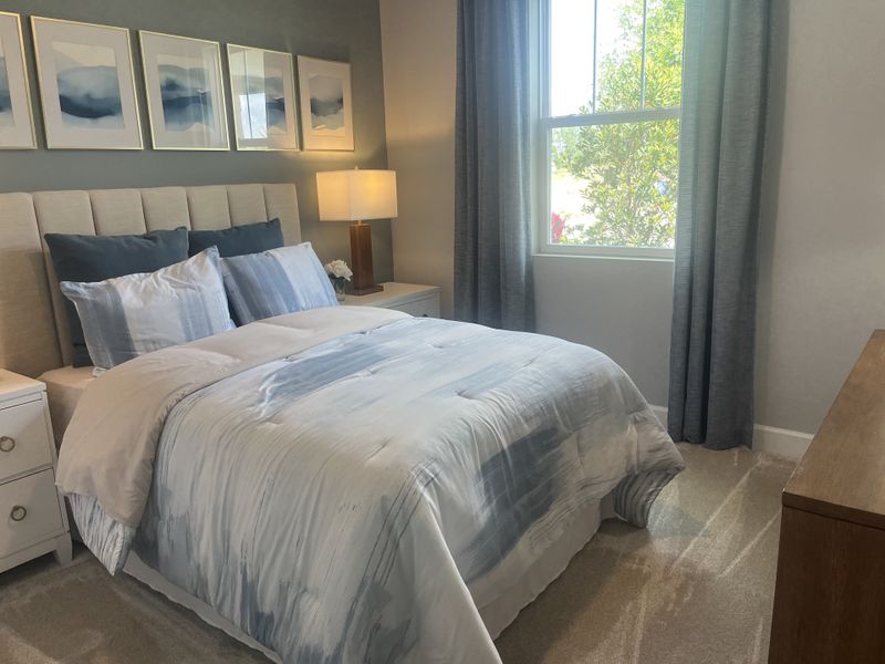 A serene bedroom with a cushioned headboard, abstract art, soft bedding, and a large window with gray curtains.