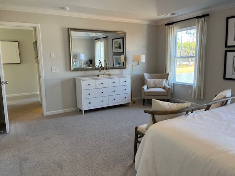 A serene bedroom with a plush carpet, elegant dresser, large mirror, and cozy seating by a sunlit window.
