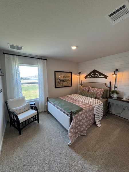 A cozy bedroom featuring a patterned bedspread, rustic wall decor, and a comfortable chair near a bright window.