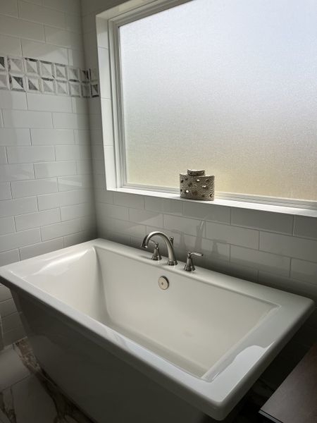 A sleek bathroom featuring a modern white tub, geometric tile accents, and a frosted window for privacy.