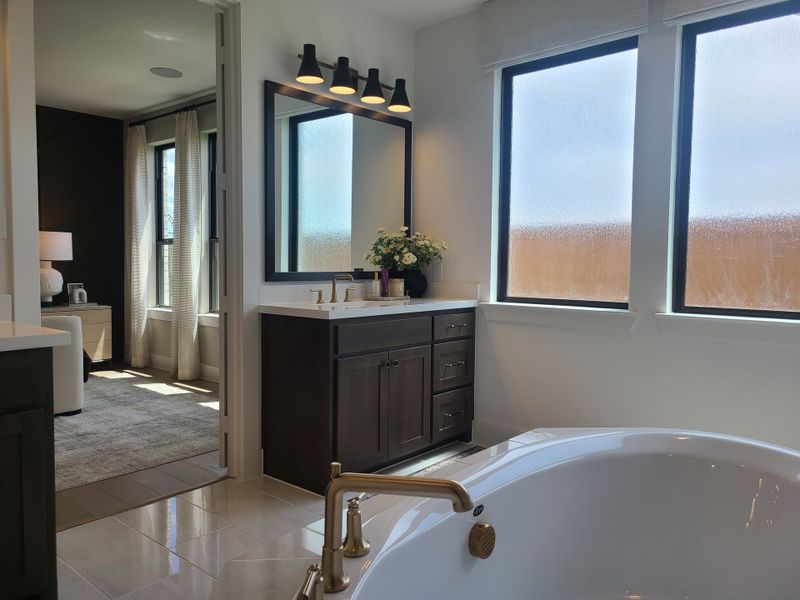 A modern bathroom featuring a freestanding tub, dark vanity, and ample natural light from large windows, leading to a cozy bedroom.