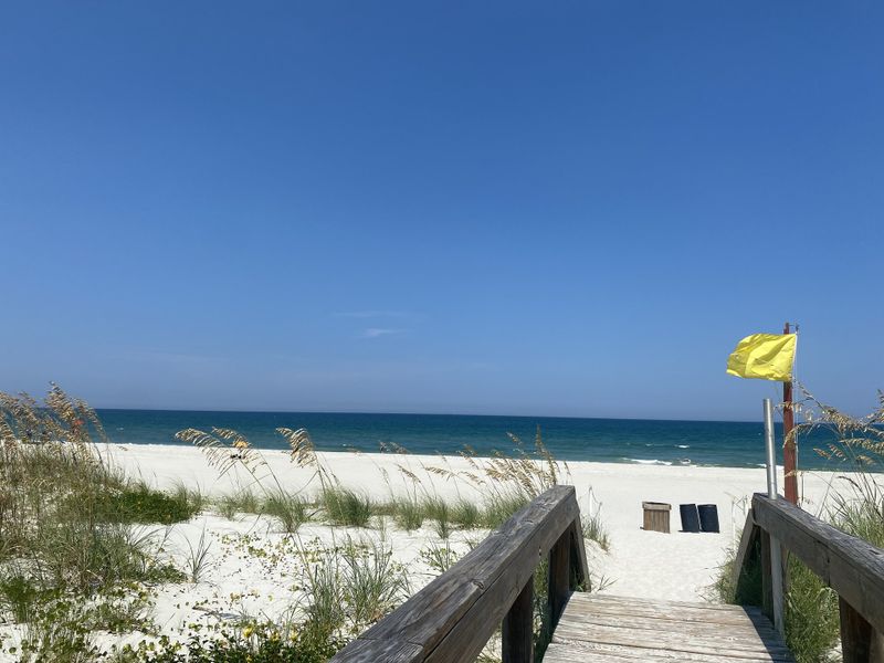 A serene beach view with a wooden pathway, white sand, and vibrant blue waters under a clear sky.